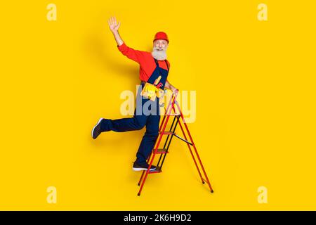 Photo pleine longueur de drôle positif vieux gars habillé uniforme général rouge hardhat levant les escaliers agitant bras isolé couleur jaune arrière-plan Banque D'Images