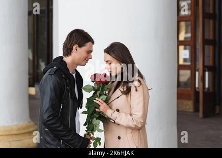 vue latérale de l'homme heureux souriant près de la jolie petite amie odeur roses près de la colonne blanche, image de stock Banque D'Images