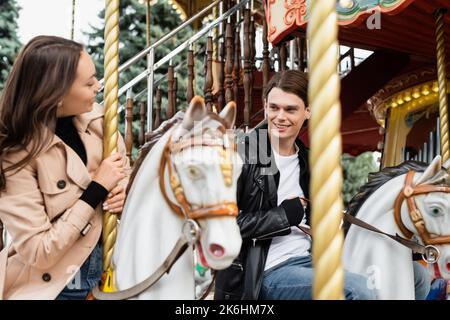 joyeux jeune homme regardant la petite amie à cheval de carrousel dans le parc d'attractions, image de stock Banque D'Images