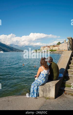 Lacs d'Italie, vue arrière d'un couple d'âge moyen en été assis ensemble sur des marches au bord du lac à Bellagio et en regardant les nageurs au lac de Côme, Italie Banque D'Images