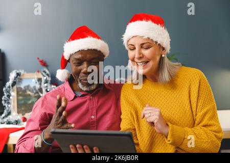 Joyeux couple senior varié en chapeau de père noël en faisant appel vidéo de noël avec une tablette Banque D'Images