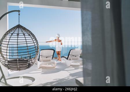 Après la douche belle femme avec cheveux et serviette de corps envelopper debout sur la terrasse de maison ouverte avec chaises longues, chaise suspendue appréciant paysage marin et bleu propre Banque D'Images