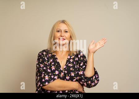 Belle femme caucasienne aux cheveux blonds et une robe à motif sombre offrant quelque chose dans une main comme un signe de générosité et d'hospitalité, isolée Banque D'Images