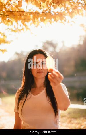 Belle jeune femme tenant la feuille jaune devant son visage tout en se tenant dans le parc contre la lumière du soleil pendant le temps ensoleillé en automne Banque D'Images