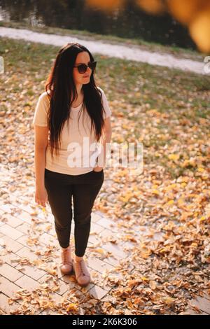 Portrait en longueur de la jeune femme heureuse et élégante en lunettes de soleil, en regardant loin en se tenant au parc par temps ensoleillé en automne Banque D'Images