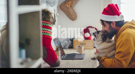 Fête de Noël à la maison avec un seul homme en vidéoconférence sur ordinateur portable qui embrasse son adorable chien. Concept de personnes appréciant le dece Banque D'Images