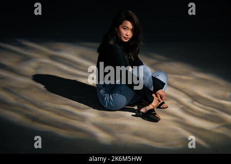 La femme retirée est assise à pieds croisés sur le sol dans une pièce sombre, un projecteur sur elle. Portrait dramatique pour la solitude, l'isolement, l'anxiété, la dépression. Banque D'Images