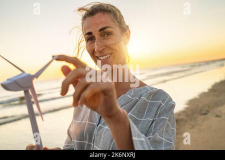 Femme souriante tenant un modèle d'éolienne au coucher du soleil sur la plage. Éolienne éolienne image du concept de la ferme d'énergie éolienne Banque D'Images