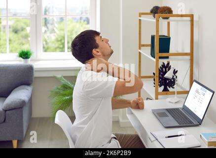 Homme fatigué et stressé assis au bureau avec ordinateur portable et tenant la main sur son cou douloureux Banque D'Images