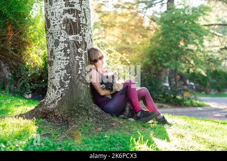 Femme blanche blonde d'âge moyen avec des lunettes se penchant contre un arbre et câliner son chien de berger catalan. Amour pour les animaux. Soin des animaux. Passion pour Banque D'Images