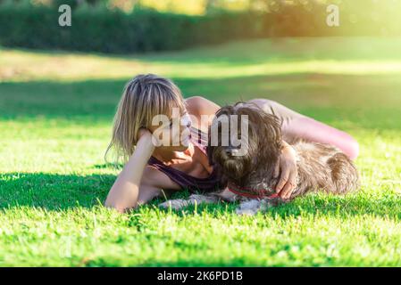 Femme blonde d'âge moyen, couchée sur l'herbe à côté de son berger catalan. Complicité et amour pour les animaux. Les animaux de compagnie apportent tranquillité et bien-être Banque D'Images