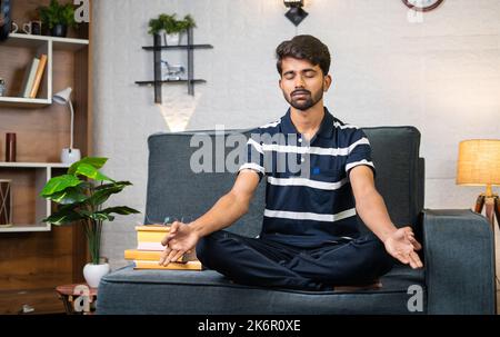 Jeune garçon indien méditant avec les yeux fermés avant de lire tout en étant assis sur un canapé à la maison - concept de soulagement de stress, la connaissance et la relaxation. Banque D'Images