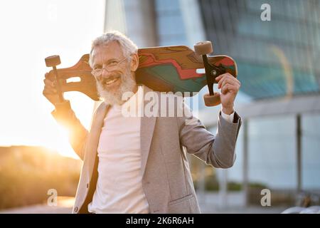 Heureux cool homme d'affaires senior homme patineur tenant le skateboard dans la ville. Banque D'Images