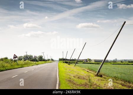 les poteaux téléphoniques se penchent du vent le long d'une route de campagne en angleterre Banque D'Images