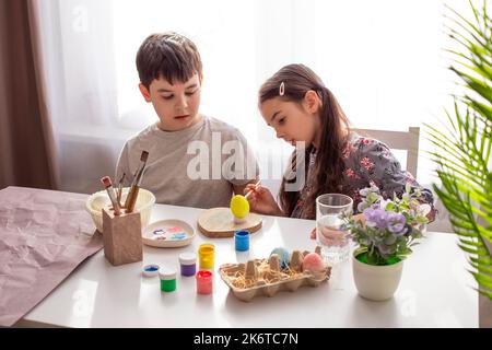 Une petite fille et un garçon brunette assis à une table blanche près de la fenêtre, peint des oeufs Banque D'Images