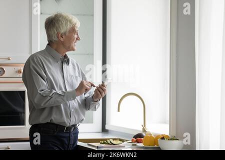 Homme plus âgé debout seul dans une cuisine moderne avec un smartphone Banque D'Images
