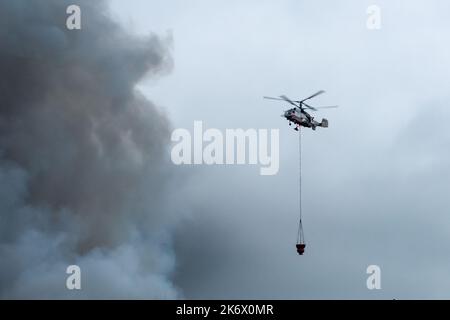 Un hélicoptère d'incendie transporte un conteneur d'eau pour éteindre un incendie dans un bâtiment de production Banque D'Images