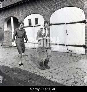 1964, une adolescente étudiante à wellington bottes et haut se réveillant à travers une ferme, Bucks, Angleterre, Royaume-Uni, derrière une femme élégamment habillée en haut, jupe, talons et portant des gants, probablement un professeur. Basé à Wendover, l'établissement d'enseignement connu sous le nom de Farmhouse School, a été établi par Quaker, Isobel Fry vers 1900 et a eu des élèves de 8 à 18 ans. Les élèves devaient se lever tôt chaque jour, car ils étaient chargés de s'occuper des animaux et des tâches générales de la ferme avant le petit déjeuner. Des cours d'école traditionnels auraient alors lieu. Banque D'Images