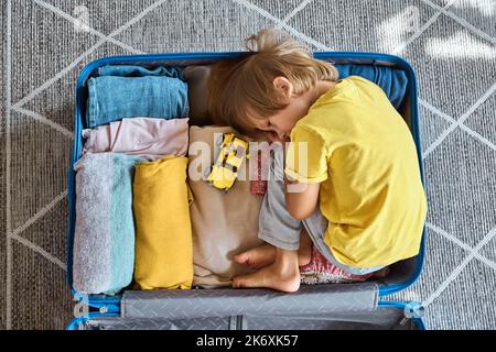 Un enfant triste se trouve dans une valise qui attend le départ et joue avec une voiture-jouet. Déménagement de la famille, vol du pays Banque D'Images