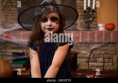 Mignon enfant fille enchantress, dans le chapeau de magicien, assis contre un foyer couvert de toile d'araignée avec livres de charme, sourires à l'appareil photo Banque D'Images