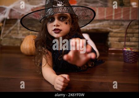 Petite fille sorcière dans un chapeau de magicien, étire sa main, regarde la caméra avec un regard effrayant. Halloween Banque D'Images