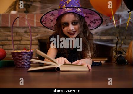 Mystérieuse petite sorcière dans un chapeau d'assistant, regarde la caméra, apprenant un livre de sort, couché contre une cheminée couverte de toile d'araignée Banque D'Images