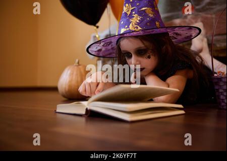 Petite fille espiègle avec maquillage Halloween vêtue de sorcière, sorcière dans le chapeau de magicien lisant un livre de sorcellerie Banque D'Images