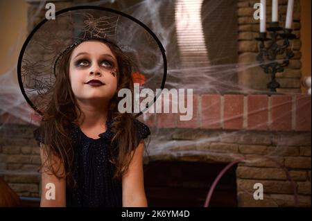 Mystérieuse petite fille en robe noire et chapeau de magicien est assis contre une cheminée couverte de toile d'araignée avec des bougies. Halloween Banque D'Images