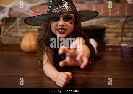 Petite fille sorcière dans un chapeau de magicien, étire sa main, regarde la caméra avec un regard effrayant. Halloween Banque D'Images