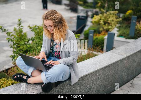 Femme à poil dur concentrée freelancer portant des lunettes dactylographiant sur l'argent portable assis sur la rue de la ville, recherchant l'Internet. Université Banque D'Images