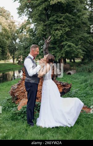 Vue latérale du couple de mariage debout près des racines de l'arbre tombé dans les doigts entrelacés du parc. Jeune femme levant une jambe. Banque D'Images