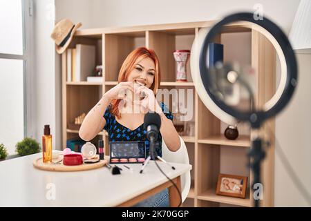 Jeune femme caucasienne appliquant maquillage faire le tutoriel souriant dans l'amour montrant le symbole de coeur et la forme avec les mains. concept romantique. Banque D'Images