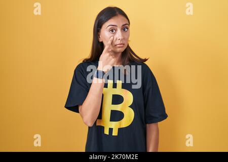 Jeune femme hispanique portant le t-shirt bitcoin pointant vers l'œil vous regardant le geste, l'expression suspecte Banque D'Images