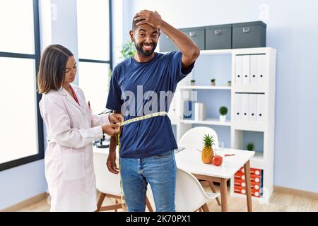 Diététiste à la clinique en utilisant un mètre ruban mesurant la taille du client stressé et frustré avec la main sur la tête, surpris et fâché visage Banque D'Images