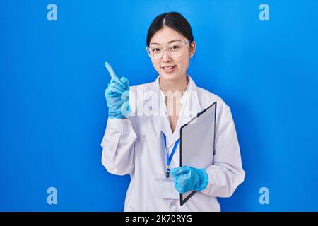 Jeune femme chinoise travaillant au laboratoire scientifique pointant du doigt vers le côté montrant une publicité, un visage sérieux et calme Banque D'Images