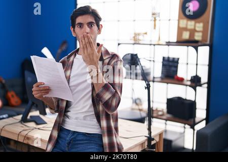 Jeune hispanique lisant la feuille de musique au studio de musique couvrant la bouche avec la main, choqué et peur de l'erreur. expression surprise Banque D'Images