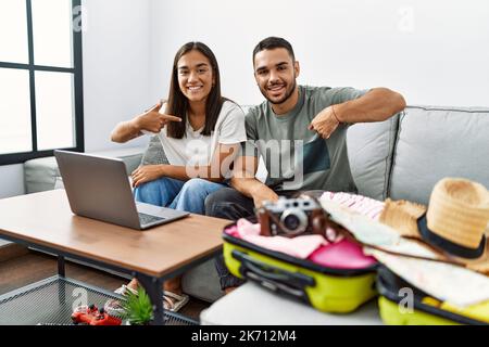 Jeune couple interracial emballant les vêtements d'été dans une valise regardant l'ordinateur portable pointant le doigt vers un auto sourire heureux et fier Banque D'Images