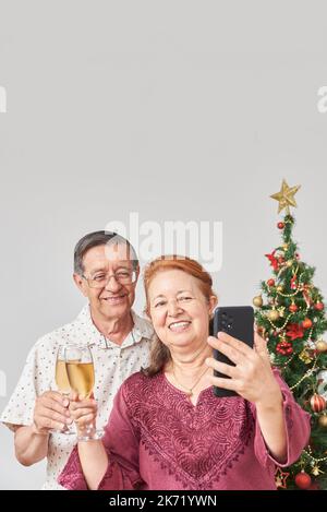 Heureux couple latin senior faisant un selfie avec un smartphone tout en toaster pendant Noël ou le nouvel an, célébrant des vacances à la maison, ayant un bon t Banque D'Images