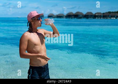 Un jeune homme boit de l'eau sur la plage avec une vue imprenable sur l'océan. Maldives Banque D'Images