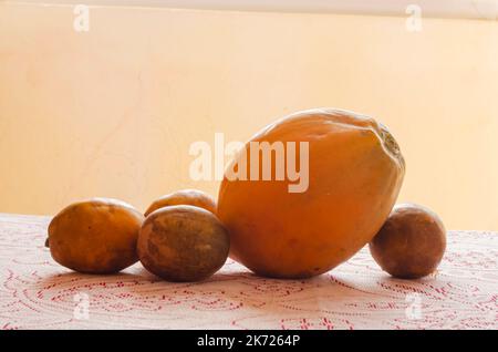 Une papaye et quatre prunes de juin sont sur une nappe en dentelle rouge et blanche. Banque D'Images