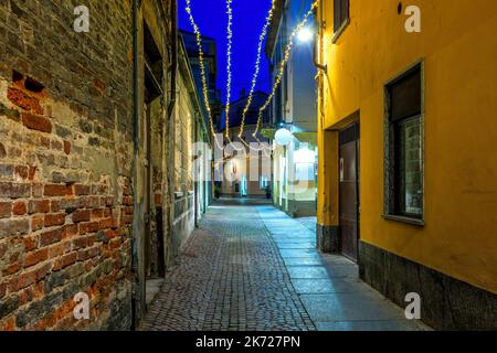 Rue pavée étroite parmi les maisons anciennes illuminées de lumières de Noël en soirée à Alba, Piémont, Italie du Nord. Banque D'Images