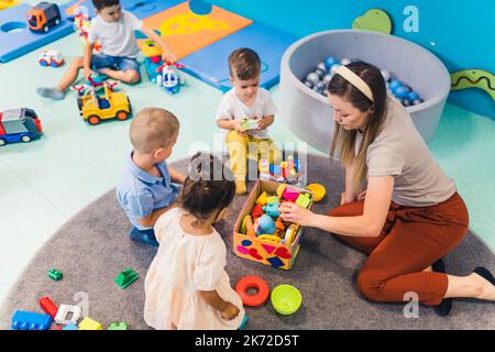 Jeune professeur caucasien assis avec ses enfants et jouant avec des jouets en plastique, jardin d'enfants et concept de jeux créatifs. Photo de haute qualité Banque D'Images