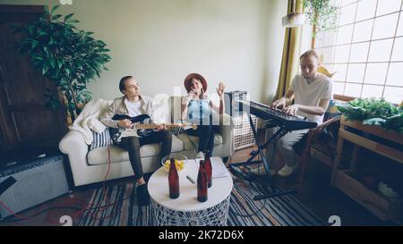 Le groupe musical répète à la maison, la femme blonde chante et les hommes jouent des instruments de musique guitare et clavier. Des bouteilles de bière et des collations sont visibles sur une table moderne. Banque D'Images