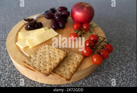 Une planche à découper au fromage avec des tranches de fromage, des tomates cerises, une pomme rouge, des raisins rouges, des craquelins, chutney picke et une cuillère à pickle, sur un travail de cuisine su Banque D'Images