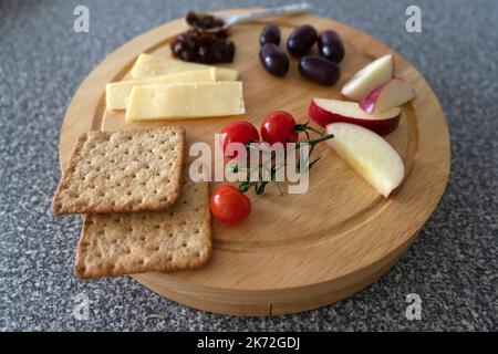 Une planche à découper au fromage avec du fromage tranché, des tomates cerises, des tranches de pomme rouge, des raisins rouges, des craquelins chutney cornichon et une cuillère à cornichons, sur une cuisine Banque D'Images