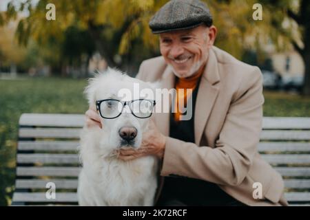 Homme senior heureux regardant l'appareil photo et embrassant son chien portant des lunettes sur le banc à l'extérieur de la ville. Banque D'Images