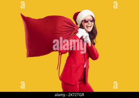 Drôle moderne Santa femme riant bruyamment portant un grand sac rouge avec des cadeaux sur ses épaules. Banque D'Images