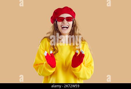 Gaie riant femme dans des lunettes rouges, chapeau et gants, chemise de siège jaune sur fond beige. Banque D'Images