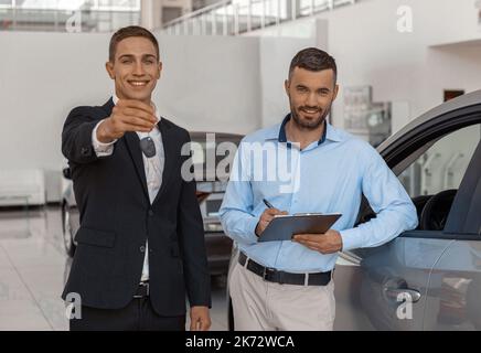 L'acheteur signe le contrat pour une nouvelle voiture en salon de l'auto tandis que le conseiller masculin montre la clé pour elle Banque D'Images