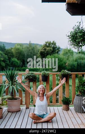 Une femme âgée assise à l'extérieur sur une terrasse en été, faisant un exercice de yoga. Banque D'Images